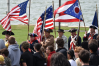 A group of people dressed in historical military uniforms carry a variety of flags, passing in front of a standing crowd.